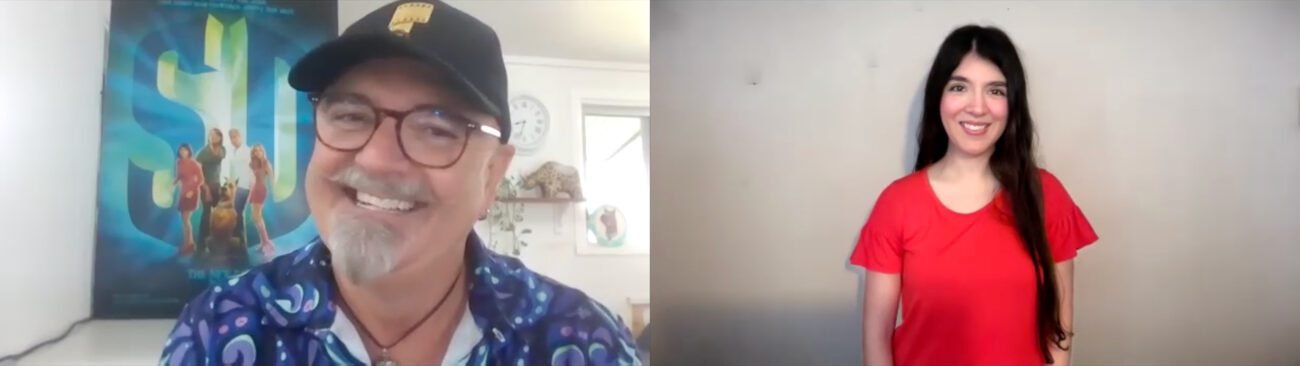 Neil Fanning, wearing a black cap and blue camp shirt with a poster for “Scooby-Doo” (2002) in the background, smiles while talking to Jamie Lee, wearing an orange shirt, in an interview.