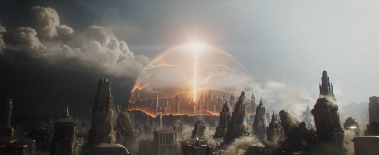 Skyscrapers of Krypton falling in close view, with a beam of orange light shining vertically into the sky during daytime in far view. The beam of light has an orange sphere surrounding it and the skyscrapers in that area of the cityscape, which remain upright. 
