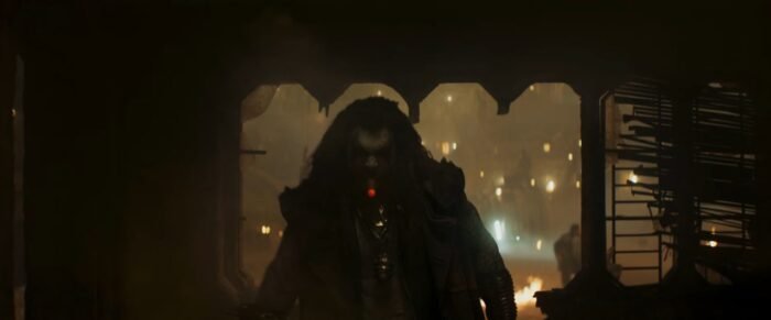 Jason Momoa as Lobo in dim light. He wears a grenade around his neck and smokes a cigar.
