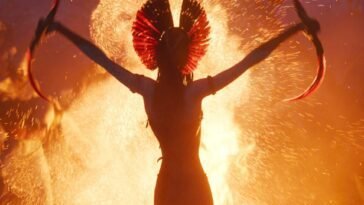 Varang stretches both of her arms wide as she holds daggers in both hands. A huge flam shoots skyward behind her.